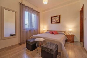 Une chambre à coucher avec un lit, deux tables de chevet, une fenêtre avec des rideaux, un tableau au mur et une petite table ronde.