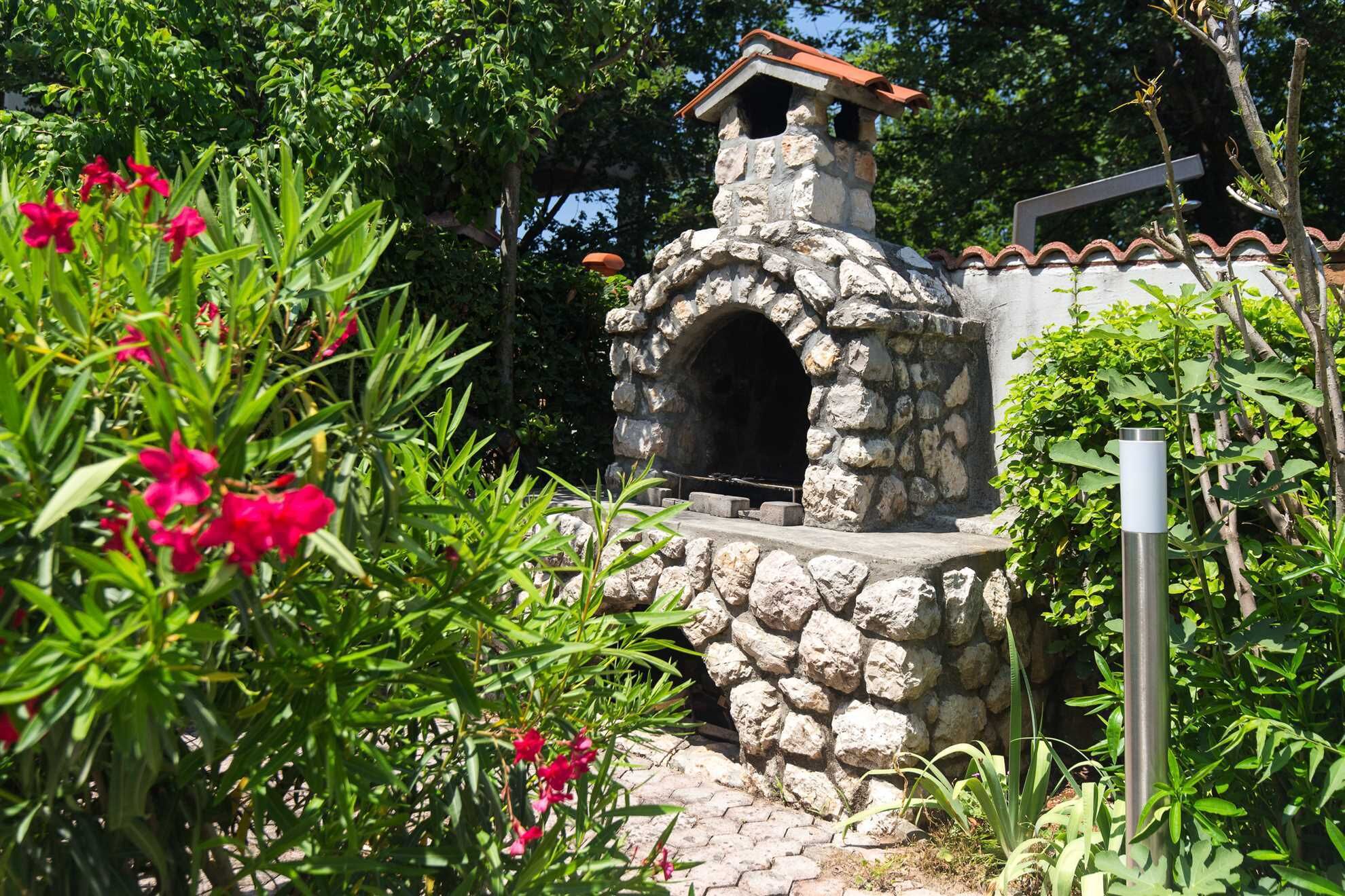 A stone-built outdoor oven with a tiled roof, surrounded by lush greenery and flowering plants.