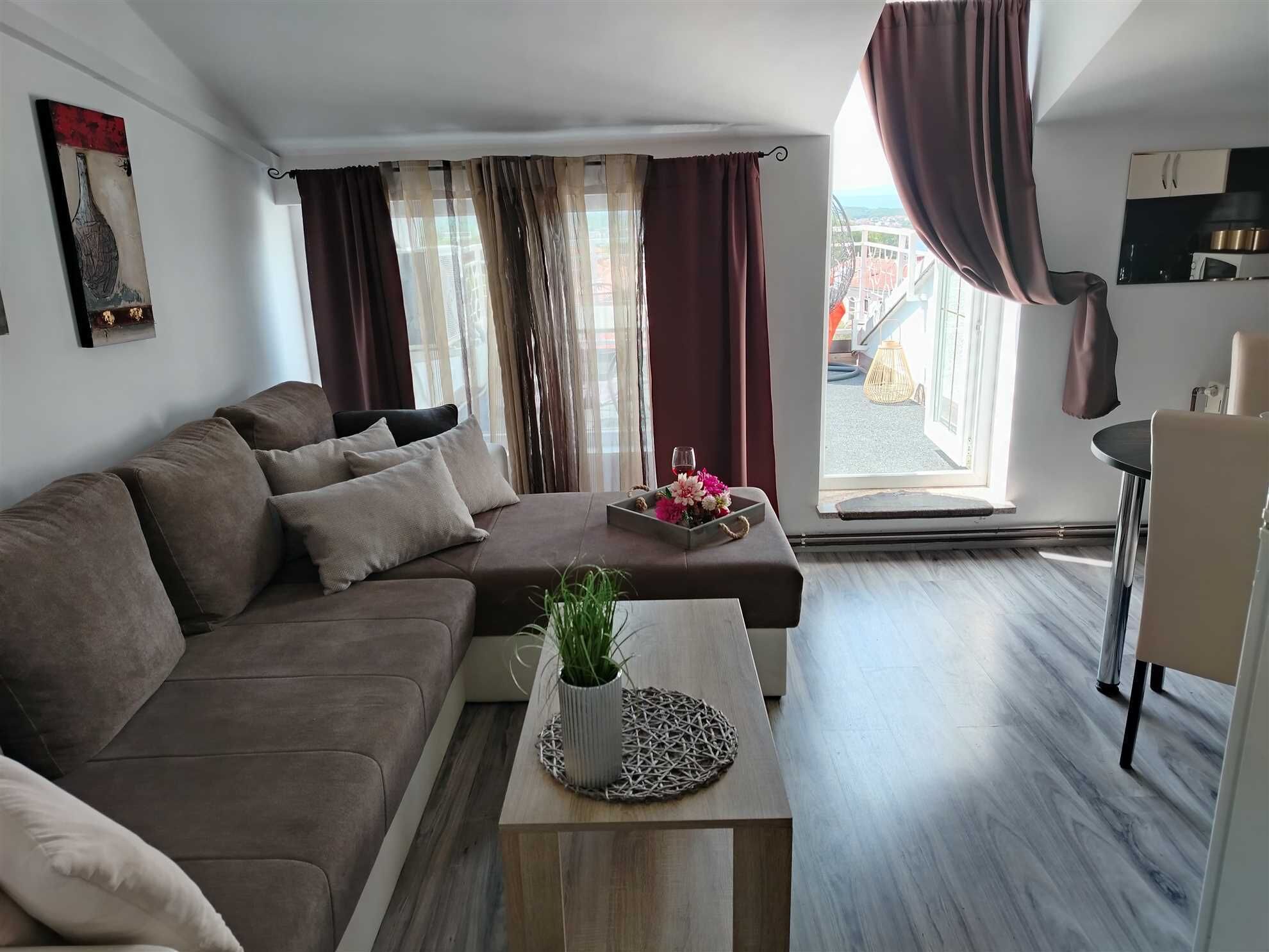 A modern living room with a sofa, a coffee table with a plant, and a view of the outside through large windows.