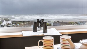 A view of the ocean through a window with binoculars and mugs on a table.