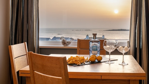 A table with a bottle of Aber Falls whisky, two glasses with ice, and a plate of snacks by a window with a view of the sea.