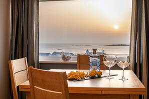 A table with a bottle of Aber Falls whisky, two glasses with ice, and a plate of snacks by a window with a view of the sea. - Bay View: 2-Bed Sea View Apartment in Rhosneigr | Managed by Anglesey Holiday Lettings (Rhosneigr)