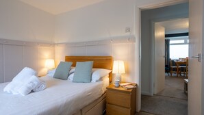A hotel room with a neatly made bed, a bedside table with a lamp, and a view of a dining area through an open door.