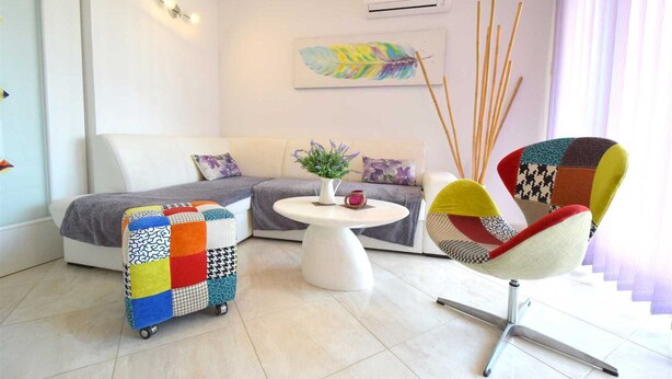 Een moderne woonkamer met een bank, een kleurrijke fauteuil, een bijzettafeltje met een bloevas en een decoratief veerme op de muur.