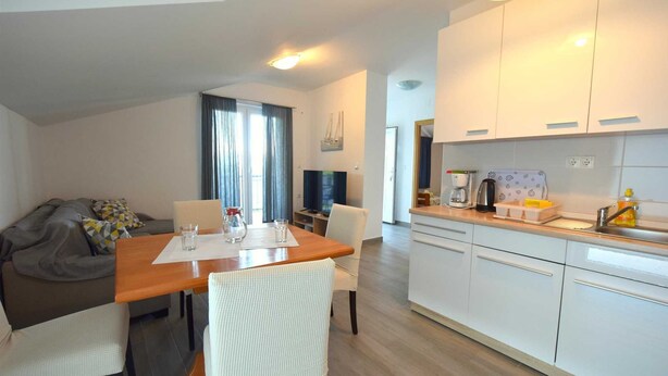 A modern kitchen with white cabinets, a dining area with a wooden table and chairs, and a living area with a sofa and cushions. - Apartment Cindrić 2 on the 2nd floor (Gabonjin)
