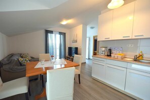 A modern kitchen with white cabinets, a dining area with a wooden table and chairs, and a living area with a sofa and cushions. - Apartment Cindrić 2 on the 2nd floor (Gabonjin)