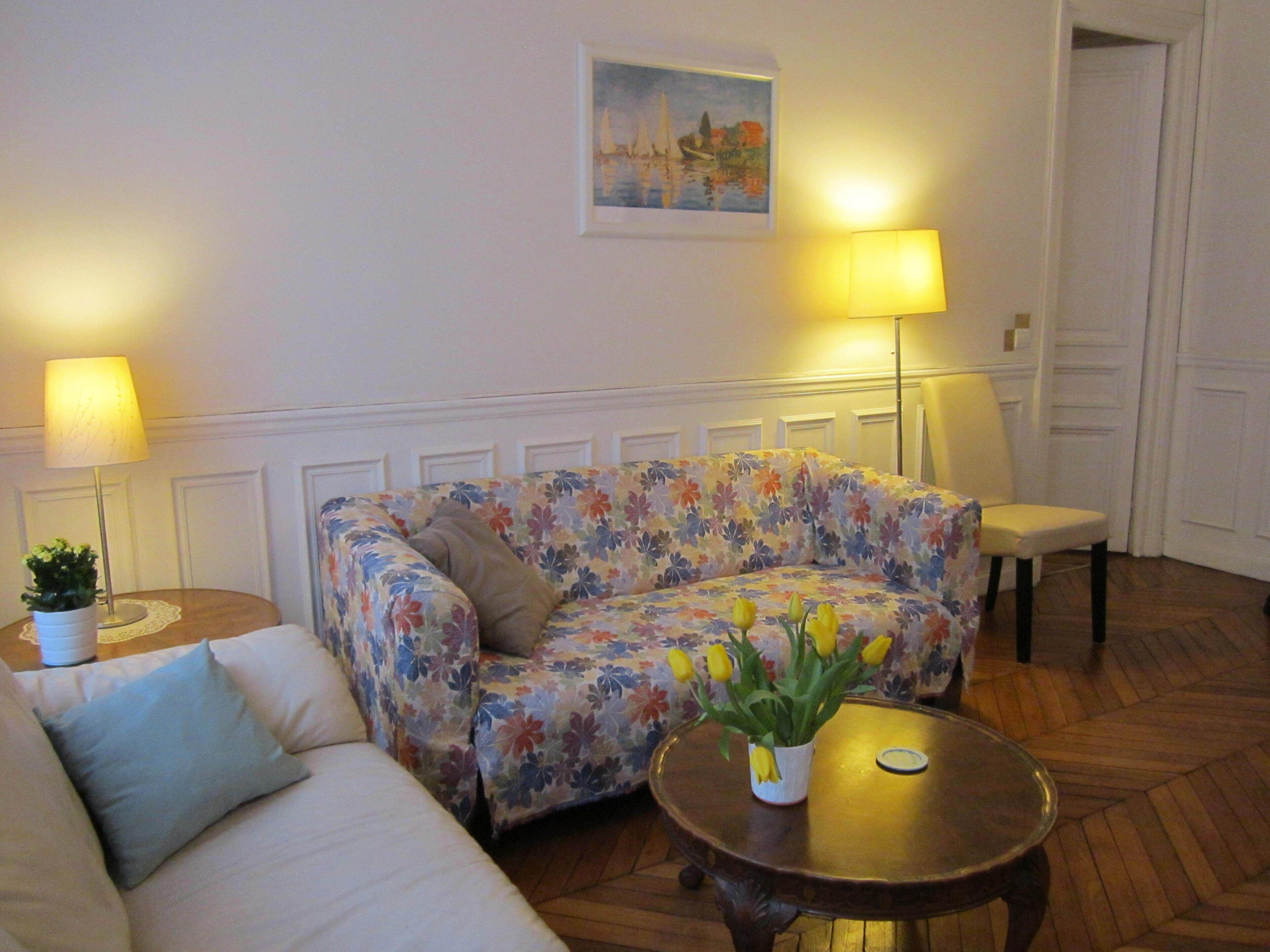 Un salon avec un canapé à motifs floraux, une petite table ronde ornée d’un vase de fleurs jaunes, et deux lampes placées de part et d’autre du canapé.