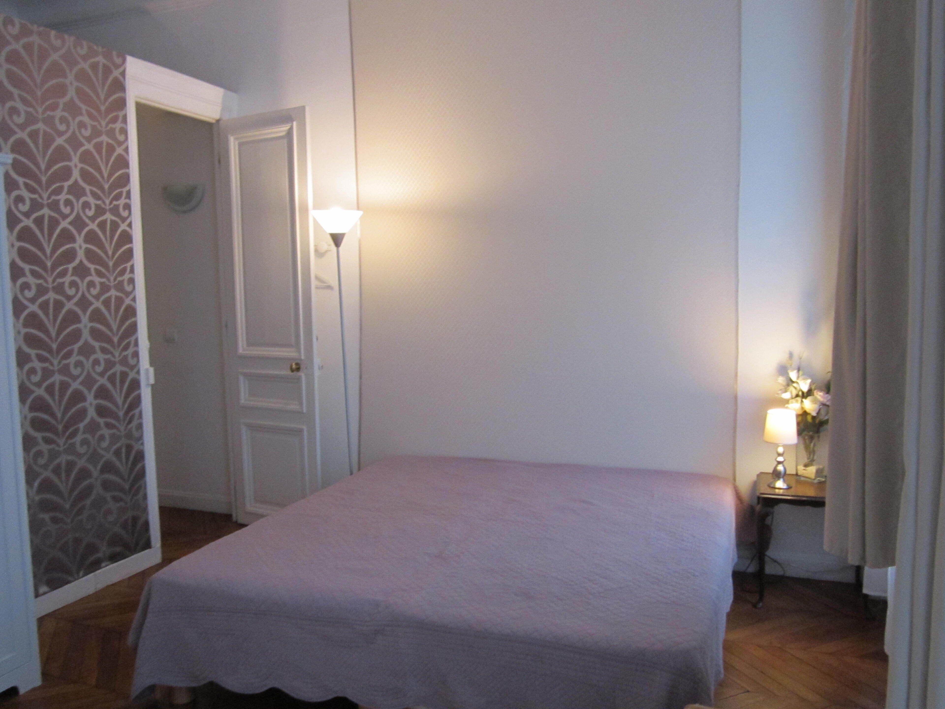 Une chambre avec un lit simple, une table de chevet avec une lampe et une porte donnant sur une autre pièce.