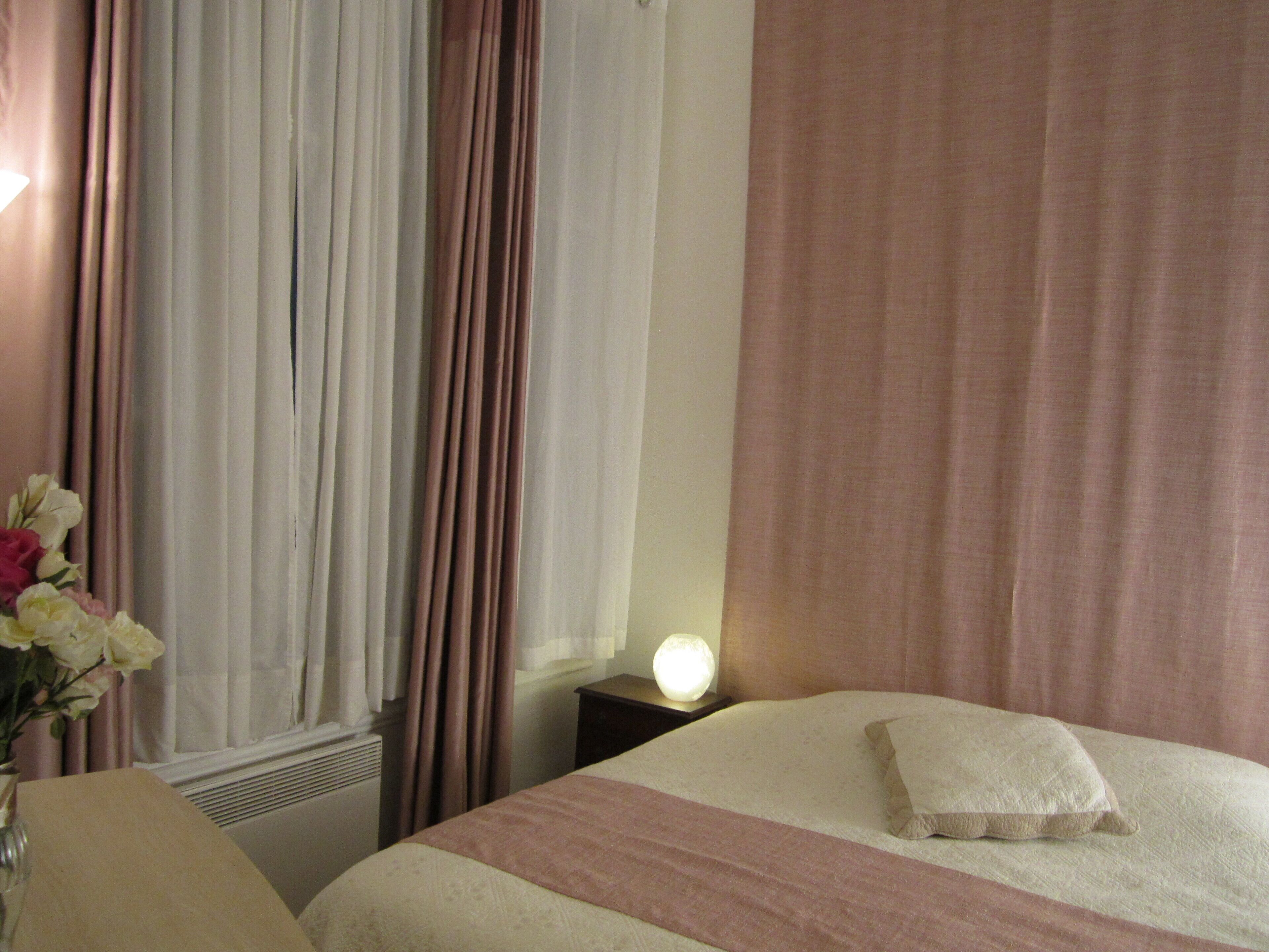 A hotel room with a bed, a nightstand with a lamp, a vase with flowers, and a window with curtains.