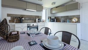 A modern kitchen with white cabinets, a checkered tablecloth, and a dining area with chairs. - Bryn Bach: Romantic 1-Bed Cottage with Hot Tub in Llanfaelog | Managed by Anglesey Holiday Lettings (Ty Croes)