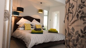 A neatly made bed with pillows and folded towels, a dark headboard, and a large mirror on the left. - Bryn Bach: Romantic 1-Bed Cottage with Hot Tub in Llanfaelog | Managed by Anglesey Holiday Lettings (Ty Croes)