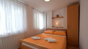 Un dormitorio con una cama, cortinas blancas, una lámpara de noche, una estantería con un jarrón y un armario de madera.