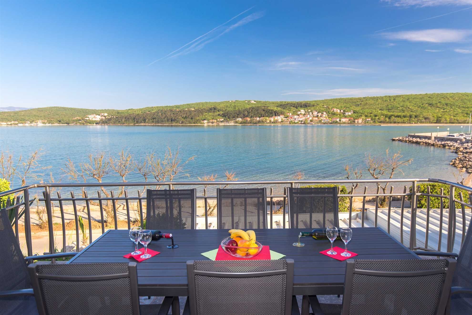 Une table avec quatre chaises, une bouteille et des verres, sur une terrasse donnant sur un lac, avec une rive visible au loin.