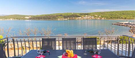 Une table avec quatre chaises, une bouteille et des verres, sur une terrasse donnant sur un lac, avec une rive visible au loin.