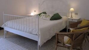 Una cama de metal blanca con un cabecero decorativo, dos mesitas de noche con lámparas, y una silla de mimbre con cojines.