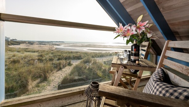 Una mesa de madera con un florero, una botella y copas, con vistas a una playa y dunas.