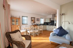 A spacious living area with a dining table, wicker chairs, a sofa, and a kitchen in the background. - Niwl Y Mor: Beachfront Rhosneigr cottage for 6 guests (Rhosneigr)