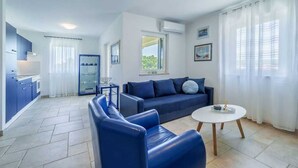 A modern living room with a sofa, a coffee table, and a kitchen area with cabinets and a refrigerator. - Apartment Jelena Punat | 300m from Beach & near Medane Beach (Punat)