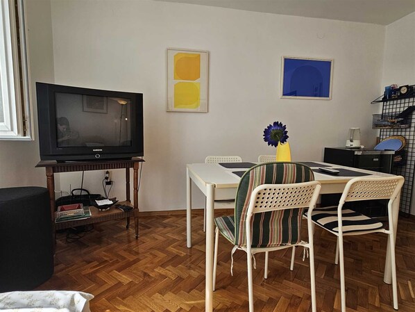 Ein kleines, möbliertes Zimmer mit Fernseher, einem Esstisch mit Stühlen sowie zwei an der Wand befestigten Kunstwerken.