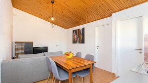 A dining area with a wooden table, grey chairs, and a grey sofa. There is a TV in the background and a door on the right.