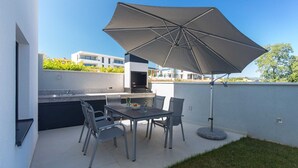 Une terrasse extérieure équipée d’un barbecue, d’une table à manger, de chaises et d’un grand parasol.
