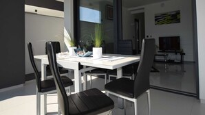 A modern dining area with a white table and black chairs, a glass partition, and a television in the background.