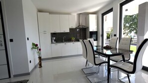 A modern kitchen with white cabinets, a dining area with a glass table and chairs, and a view of the outdoors through large windows.