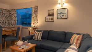 A cozy living room with a dark blue sofa, a wooden coffee table, and a dining area with a table and chairs. There are two wall-mounted lamps and framed pictures on the wall. - Marimar : Charming 3‑Bed Pet-Friendly Bungalow Steps from Rhosneigr Beach | Managed by Anglesey Holi (Rhosneigr)