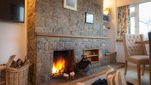 A cozy living room with a stone fireplace, a comfortable armchair, and a television on the wall. - Marimar : Charming 3‑Bed Pet-Friendly Bungalow Steps from Rhosneigr Beach | Managed by Anglesey Holi (Rhosneigr)