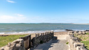 A coastal pathway leading to a beach with a stone wall on one side and a clear view of the sea and distant land. - Marimar : Charming 3‑Bed Pet-Friendly Bungalow Steps from Rhosneigr Beach | Managed by Anglesey Holi (Rhosneigr)