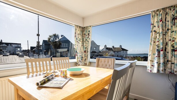 A dining area with a wooden table, chairs, and a view of the sea and houses through large windows. - Marimar : Charming 3‑Bed Pet-Friendly Bungalow Steps from Rhosneigr Beach | Managed by Anglesey Holi (Rhosneigr)
