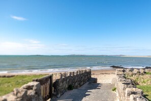 Ein Küstenweg, der zu einem Strand führt, mit einer Steinmauer an einer Seite und freiem Blick auf das Meer und das entfernte Land.