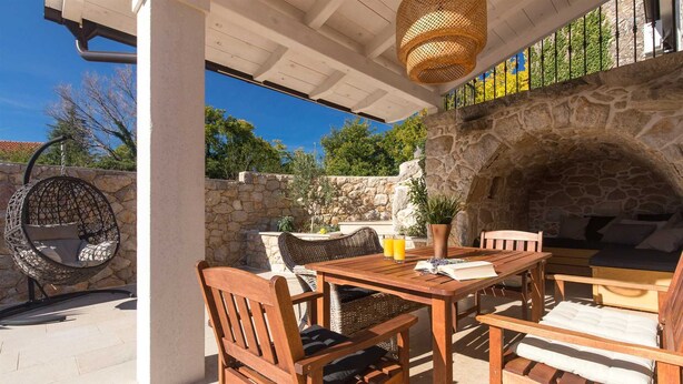 An outdoor patio with a stone wall, a hanging chair, a wooden table with chairs, and a stone fireplace.