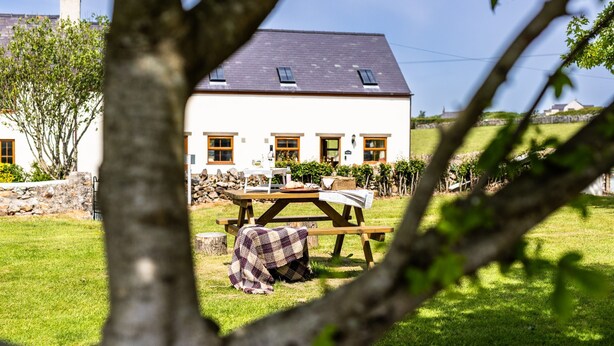 A white house with a dark roof, a picnic table, and a stone wall in the background.