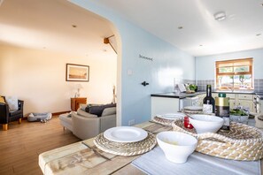 A modern kitchen with a dining area, featuring a wooden table set for a meal, a kitchen counter with appliances, and a view of the outdoors. - The Granary: A two bed, pet-friendly holiday cottage (Ty Croes)