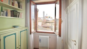 A room with a window overlooking a cityscape, a radiator, and a bookshelf filled with books. - TORRE DEL CAMPO - Two Bedroom Apartment, Sleeps 2 (Siena)