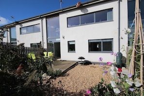 A modern two-story residential building with a wooden deck, outdoor seating, and a garden with flowers.