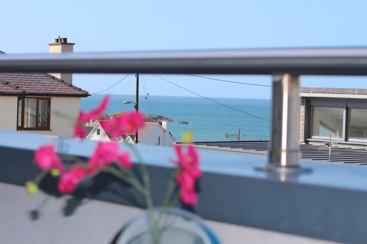 En balkong med utsikt över havet, en byggnad med ett tak täckt av tegelpannor och en blomsteruppsättning.