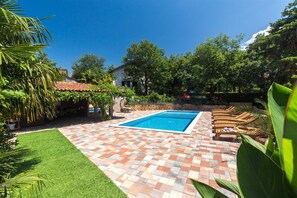 A rectangular swimming pool with a tiled deck, surrounded by greenery and a house in the background.