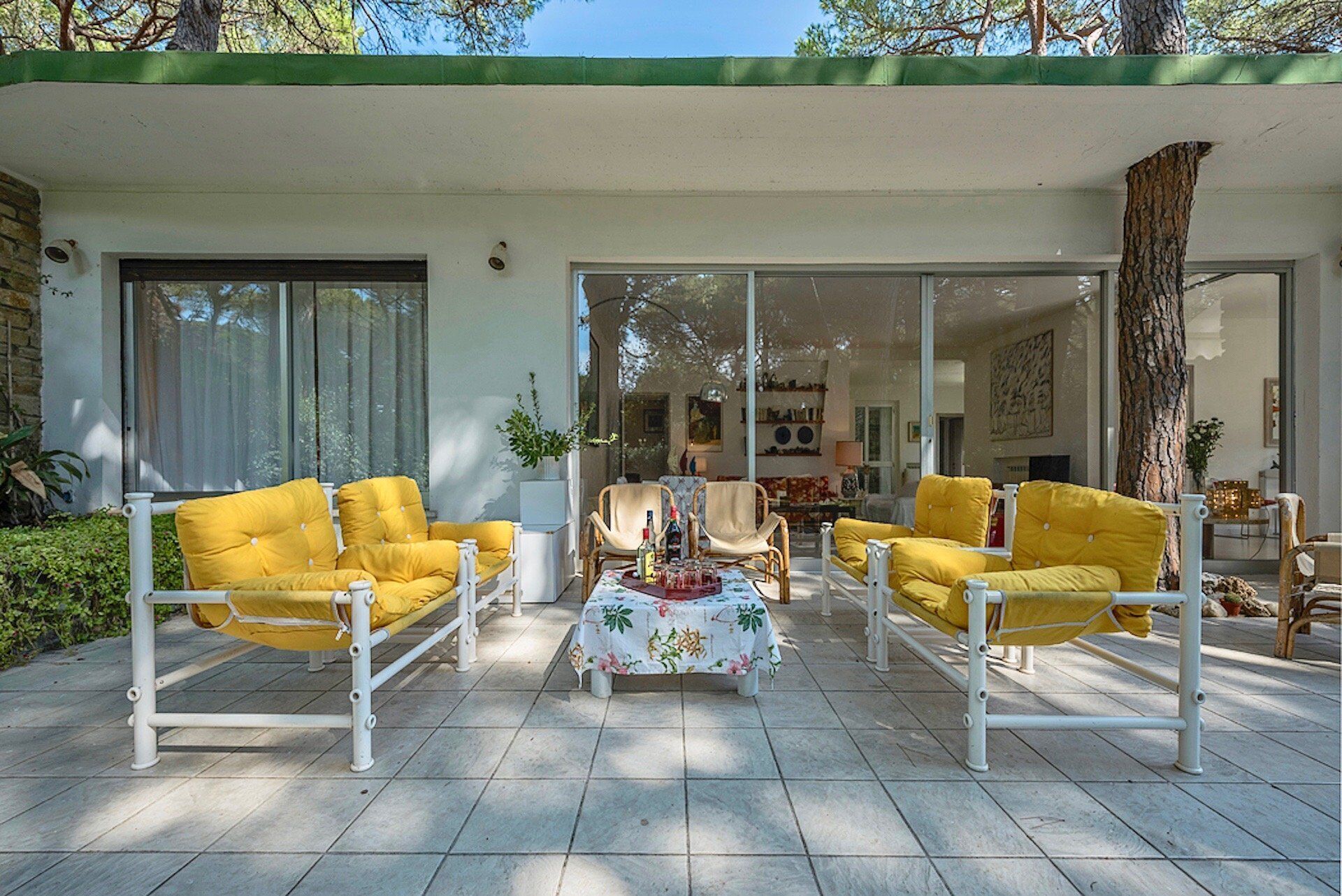 Ein überdachter Patio-Bereich mit gelb gepolsterten Stühlen und einem Tisch, umgeben von Glastüren, die zu einem Wohnzimmer führen.