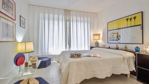 Habitación de hotel con cama, escritorio con lámpara, silla, ventana con cortinas y obras de arte en las paredes.