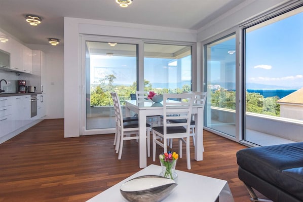 Enjoy a room with a balcony offering a scenic view of the sea and greenery, featuring a dining table, sofa, and kitchen area.