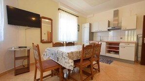 A compact kitchen with white cabinets, a built-in oven, and a microwave. There is a dining table with four chairs and a TV mounted on the wall.