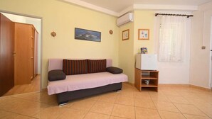 A small living room with a sofa, a TV, and a refrigerator. There is a door leading to another room and a window with curtains.