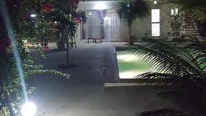 Vista nocturna de un edificio de dos pisos con balcón, rodeado de árboles y una piscina pequeña.