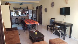 Una sala de estar con comedor, una cocina al fondo, un televisor sobre un mueble bajo y un ventilador sobre una mesa.