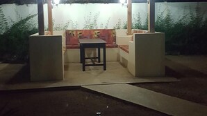 Un área de estar al aire libre con una mesa y bancos bajo un techo de paja, rodeada por un muro bajo y plantas.