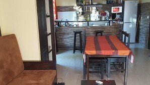 Una sala de estar con un sofá, otomanas y una mesa de centro. Al fondo, se observa una cocina con un refrigerador, microondas y diversos utensilios.