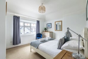 A bedroom with a bed, a desk, a chair, a dresser, a lamp, a window with curtains, and a hanging light fixture.