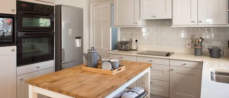 A modern kitchen with white cabinetry, a wooden table, a built-in oven, a refrigerator, a toaster, and a kettle.